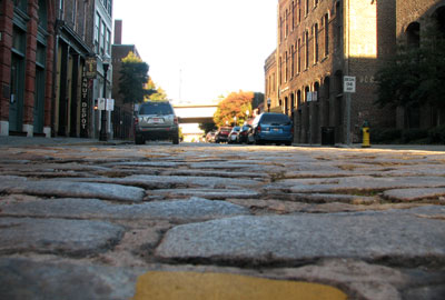 Photo of Morris Ave's cobblestone street from the level of the stones themselve. Parked cars and buildings are visible down the street.